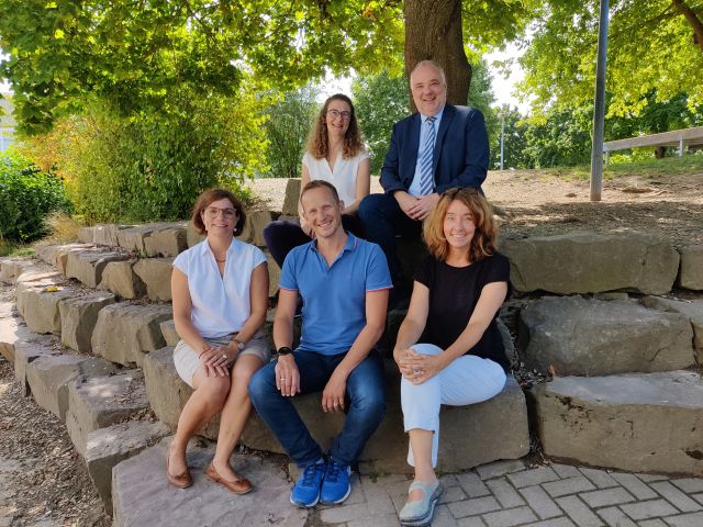Das Bild zeigt eine Gruppe von fünf Personen, die auf einer steinernen Sitzgelegenheit im Freien sitzen. Vordere Reihe: In der Mitte sitzt ein Mann mit kurzen Haaren, der ein blaues T-Shirt trägt und lächelt. Neben ihm sitzt eine Frau mit braunen, lockigen Haaren in einem weißen Oberteil. Hintere Reihe: Hinter ihnen steht ein Mann in einem Anzug mit Krawatte, der ebenfalls lächelt. Neben ihm steht eine Frau mit langen, braunen Haaren in einem weißen Oberteil. Die letzte Person in der Reihe ist eine Frau mit lockigen Haaren, die ein schwarzes Oberteil trägt. Im Hintergrund sind Bäume und eine grüne Umgebung sichtbar, was eine freundliche Atmosphäre schafft.