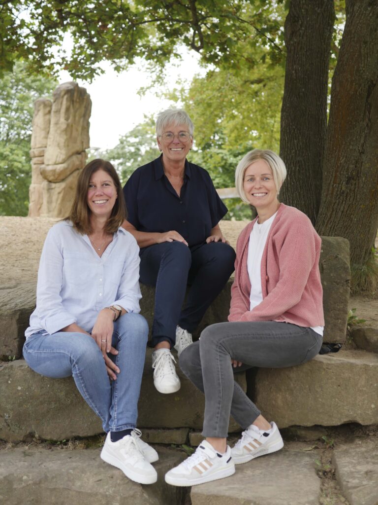 Das Bild zeigt drei Frauen, die auf einer steinernen Sitzgelegenheit im Freien sitzen. Links: Eine Frau mit langen, braunen Haaren trägt ein hellblaues Hemd und Jeans. Sie lächelt in die Kamera. Mitte: Eine Frau mit kurzen, grauen Haaren und Brille trägt ein schwarzes Oberteil und sitzt auf einem Stein. Sie lächelt ebenfalls. Rechts: Eine Frau mit kurzen, blonden Haaren trägt einen rosa Cardigan und eine weiße Bluse. Sie sitzt ebenfalls auf der Steinbank und lächelt. Im Hintergrund sind Bäume und eine natürliche Umgebung sichtbar, sowie eine große steinerne Skulptur. Die Atmosphäre wirkt freundlich und entspannt.