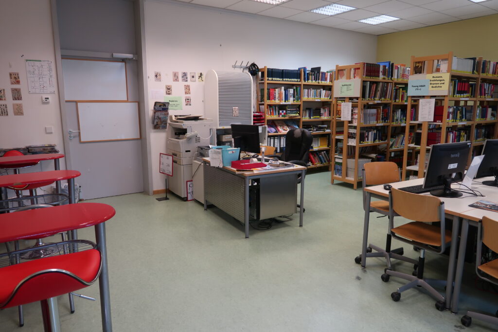 Das Bild zeigt einen Raum, der wie eine Bibliothek eingerichtet ist. Auf der rechten Seite stehen Bücherregale, die mit vielen Büchern gefüllt sind. Davor befinden sich mehrere Schreibtische mit Computern und Stühlen. Auf der linken Seite des Raumes ist eine Tür zu sehen. Davor stehen mehrere hohe Tische mit einer Reihe roter Barhocker. In der Mitte des Raumes steht ein großer Schreibtisch mit einem Computerbildschirm und verschiedenen Büroartikeln darauf. Dahinter, an der Wand, befindet sich ein Drucker oder Kopierer. An den Wänden hängen kleine Poster oder Bilder. Der Boden ist hellgrün, und die Decke ist mit Leuchtstofflampen ausgestattet.