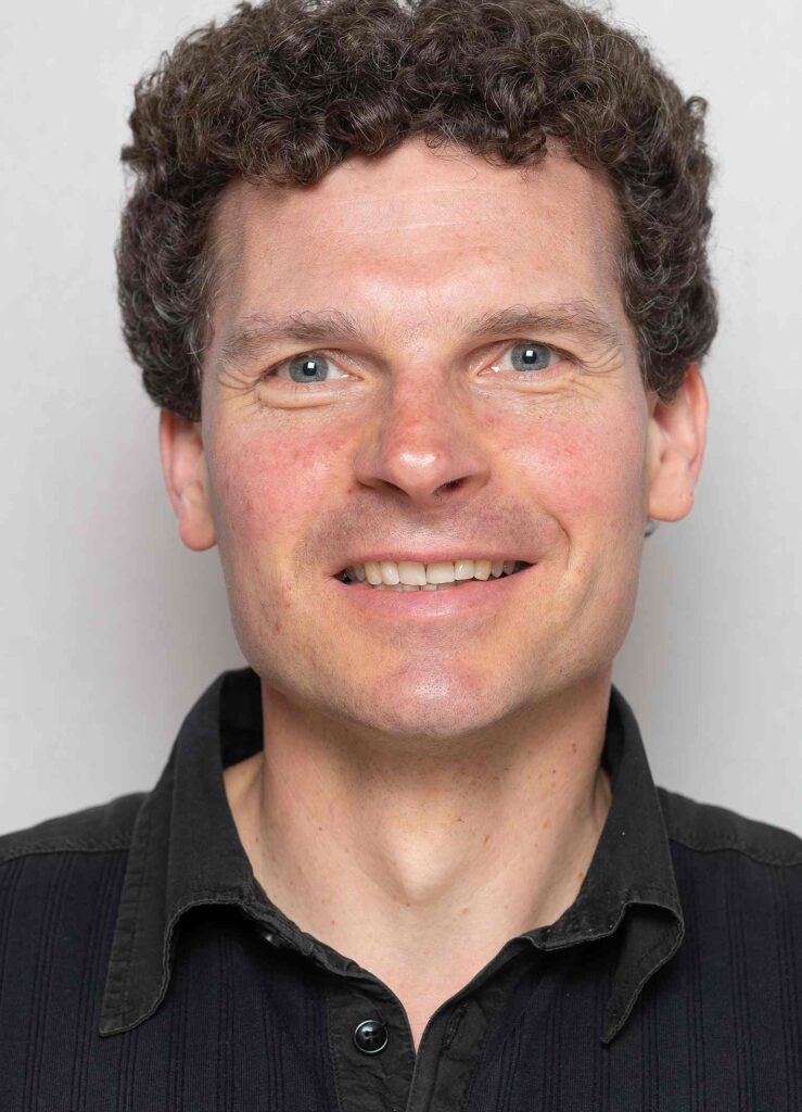 Das Bild zeigt einen Mann mit kurzem, lockigem Haar, das an den Seiten etwas länger ist. Er hat einen freundlichen Gesichtsausdruck und trägt ein schwarzes Hemd. Der Hintergrund ist hell und unauffällig, was den Fokus auf das Gesicht des Mannes lenkt.